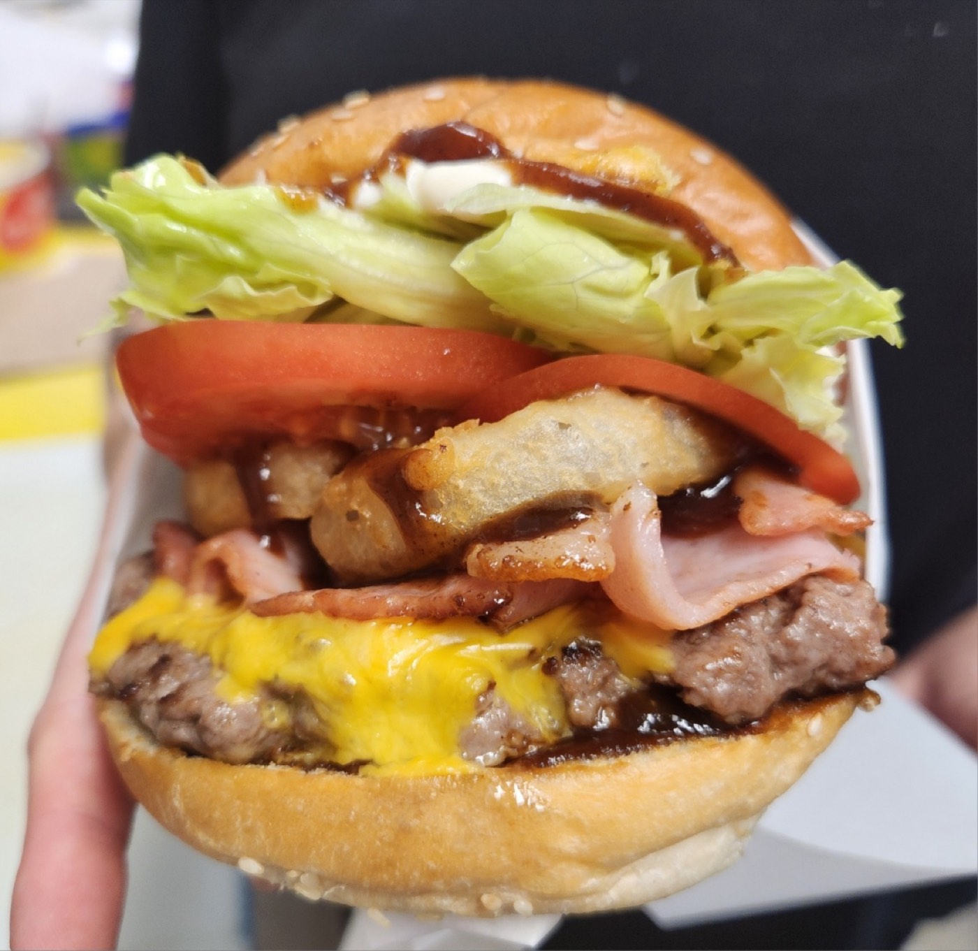 A loaded burger held in a hand, with a beef patty, melted cheese, bacon, tomato, lettuce and barbecue sauce in a glossy bun