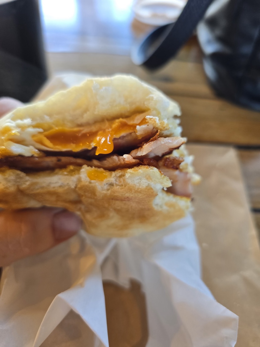 An egg and bacon roll held in a hand over paper, with a bright runny yolk and crispy bacon in a soft bun