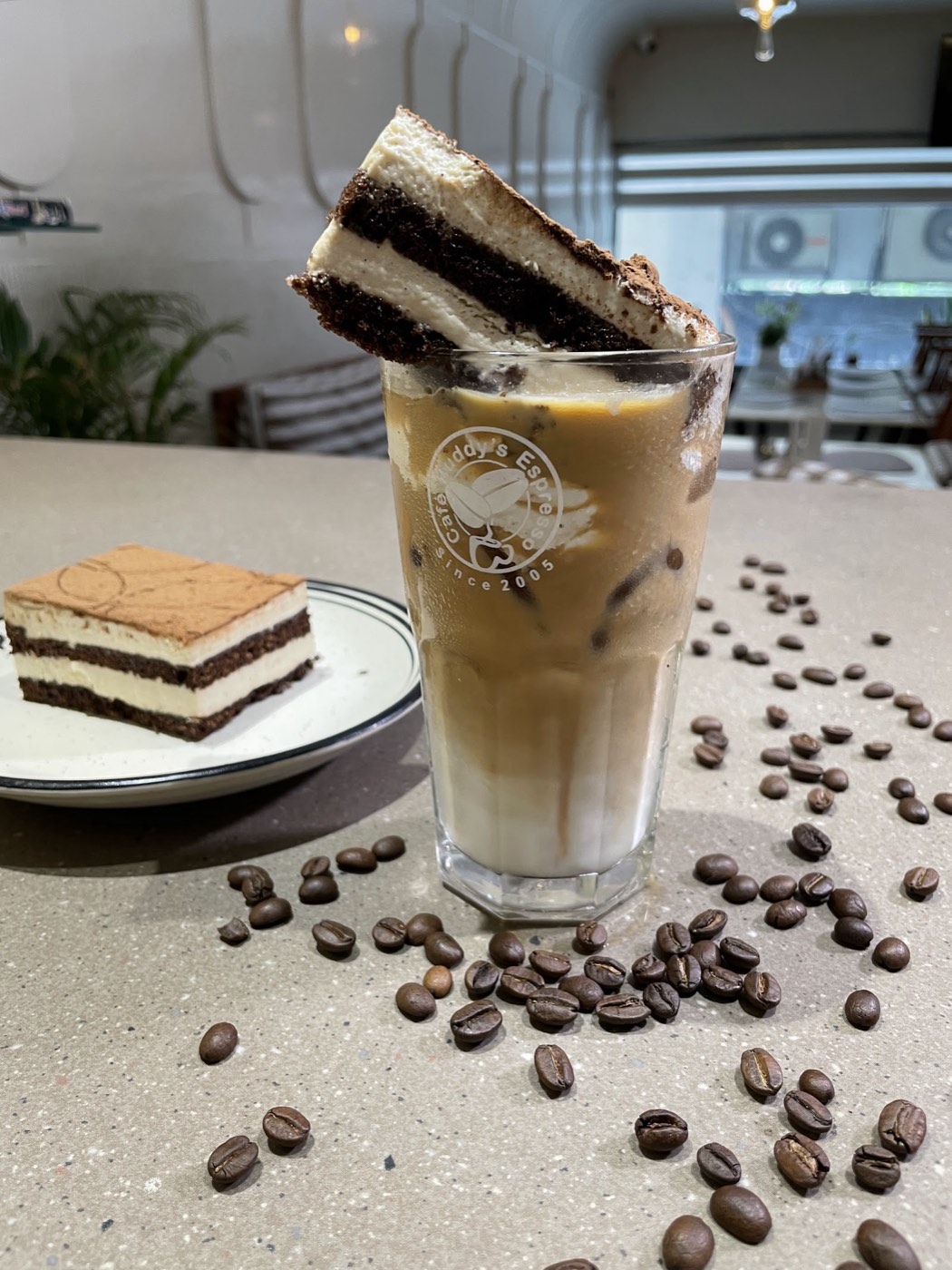 An iced coffee in a glass with a layered cake slice balanced on the rim, beside a plated slice and scattered coffee beans