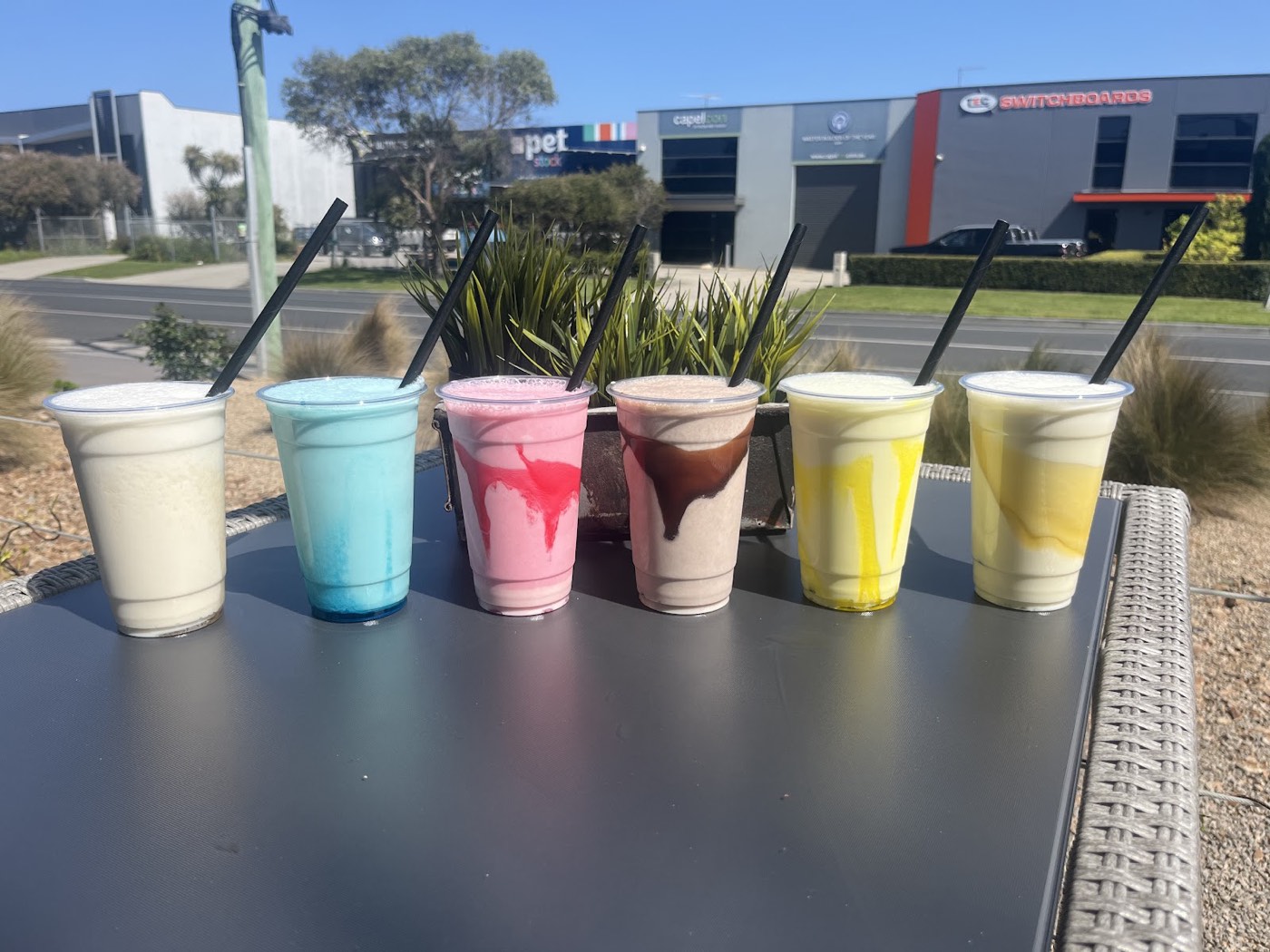 Six milkshakes in cups with black straws, in white, blue, pink, chocolate and two shades of yellow, lined up on an outdoor table