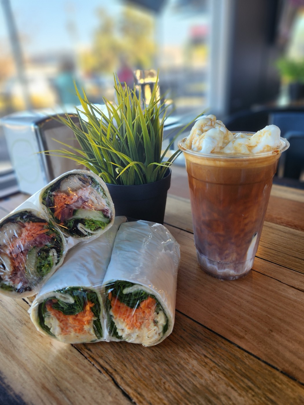 Two halved wraps filled with salad and carrot beside an iced coffee topped with cream, on a timber table